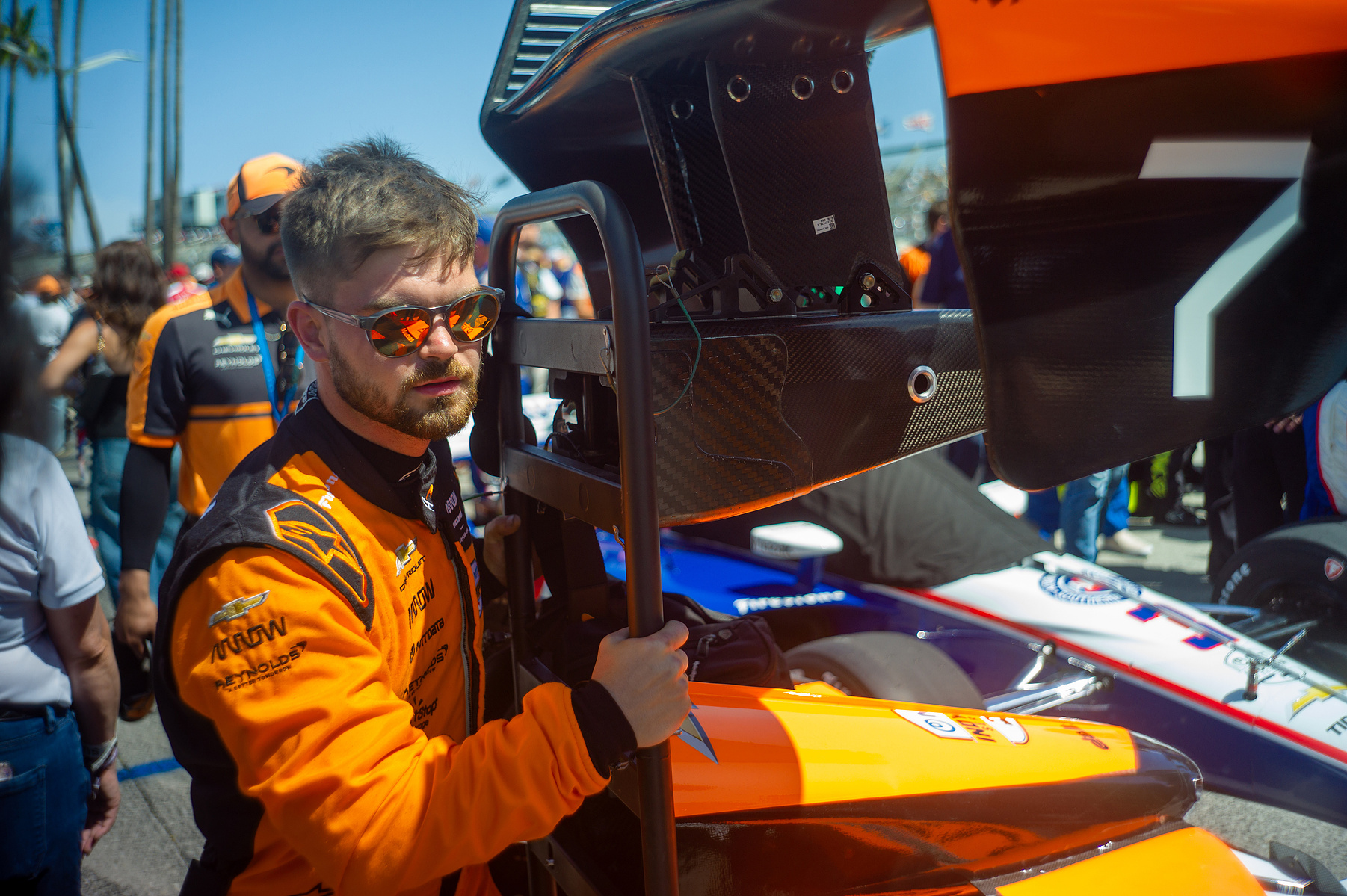 A crew member in orange carries a racing-car body panel in the Long Beach paddock.