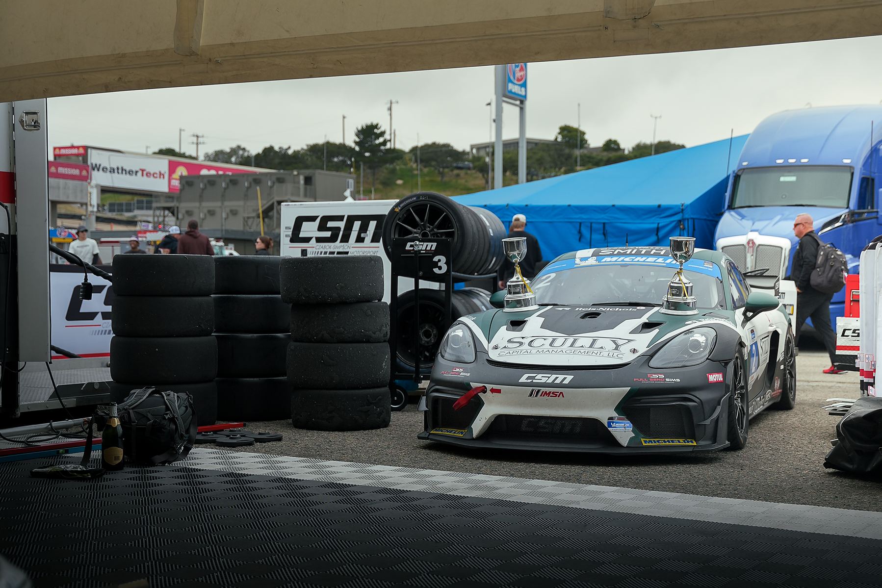 A race car waits beside tire stacks and team transporters in the Laguna Seca paddock.
