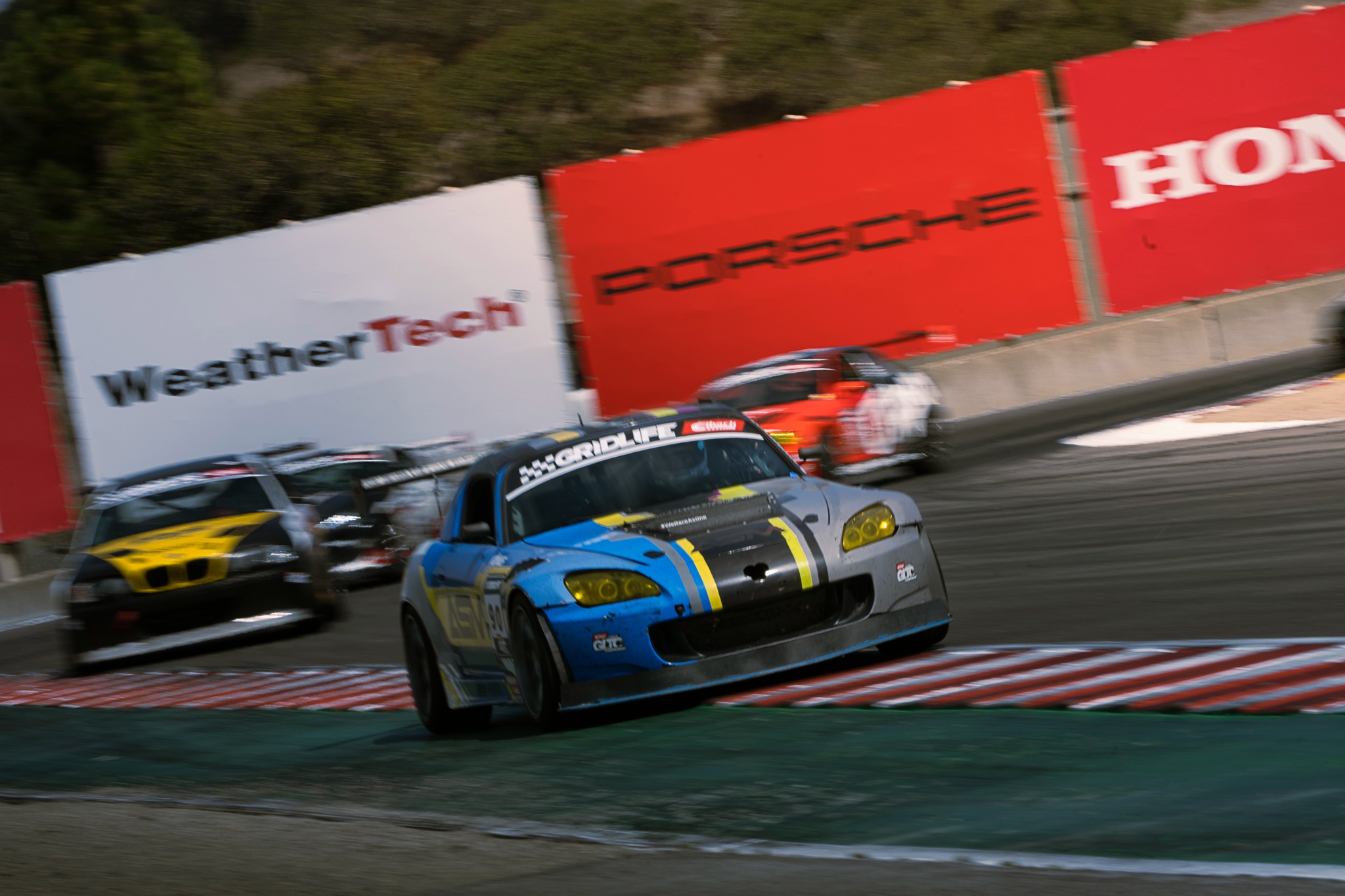 A blue and yellow race car leads a group through a Laguna Seca corner.