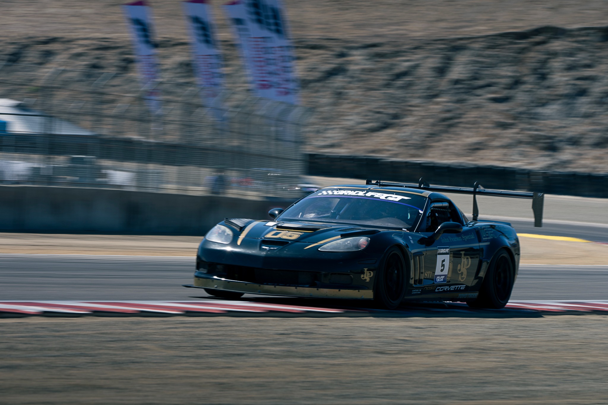 A dark sports car passes trackside fencing at Laguna Seca.